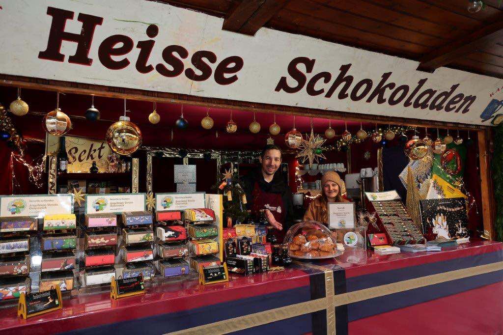 A festive chocolate stand with two vendors, colorful chocolate bars displayed, and a sign reading "Heisse Schokoladen" above.