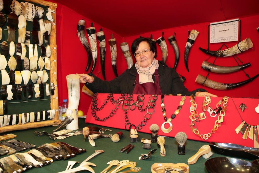 A vendor in a red booth displays horn crafts, including necklaces, combs, and drinking horns, with a variety of handcrafted items on the table.