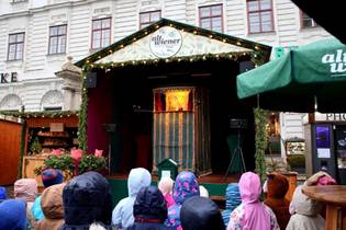 An outdoor puppet show on a stage with a festive canopy. Children in colorful hooded raincoats watch intently. The scene is lively and engaging.