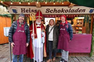 A group of four people stands in front of a festive hot chocolate stall. Two wear aprons, one is dressed as a bishop, and they all smile warmly.