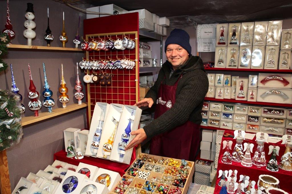 A vendor in a beanie and apron smiles while displaying Christmas ornaments in a festive market stall filled with colorful decorations.