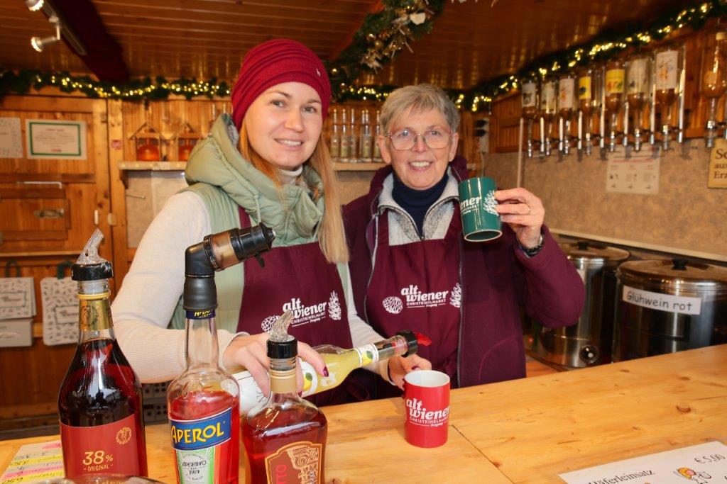 Two women in aprons serve drinks at a festive market stall, surrounded by bottles and holiday decorations.