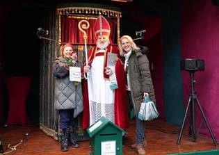 Three people stand on stage; the center figure is dressed as a traditional St. Nicholas, holding a pastoral staff. Festive, joyful atmosphere.