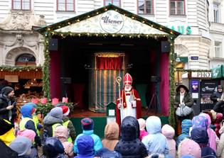 A festive scene at a Christmas market with a man dressed as St. Nicholas on a decorated stage, surrounded by children in colorful winter clothing.