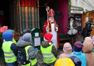 A person dressed as Saint Nicholas, in a red robe and hat, stands on a stage. They engage with attentive children in bright jackets, conveying a festive atmosphere.
