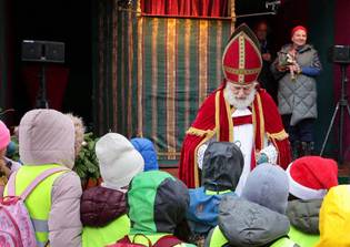 A person dressed as a traditional holiday figure, wearing a red robe and mitre, stands before a group of children in winter clothing. The mood is festive.