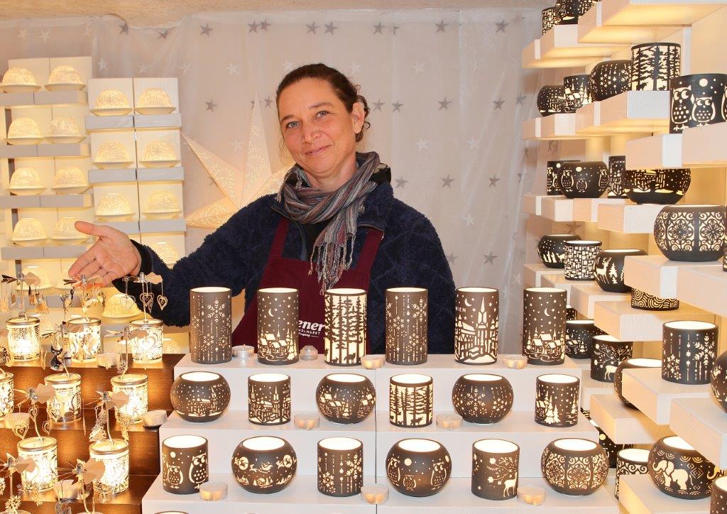A woman stands behind a display of intricately designed candle holders at a market stall, surrounded by glowing lights.