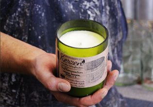A person holds a green glass candle labeled "Weinviertler Kerzen," featuring a white wax interior and a rustic, textured background.
