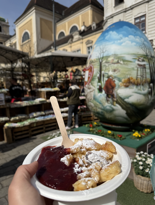 A bowl of Kaiserschmarrn with berry sauce is held in the foreground at an outdoor market with a large, decorated egg in the background.