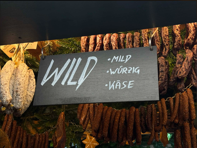 A sign labeled "WILD" with options "mild," "würzig," and "käse" hangs above clusters of hanging sausages. Background features greenery, evoking a rustic market feel.