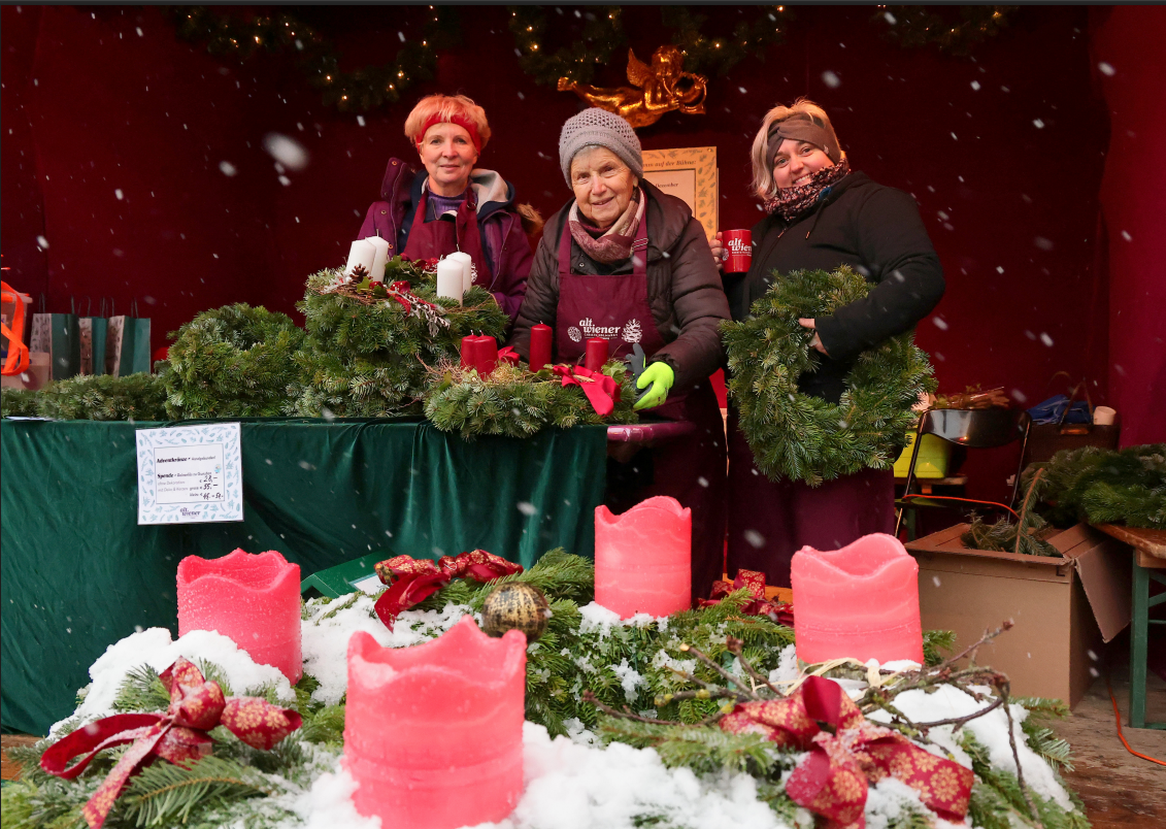 Three people stand at a festive market stall, smiling and holding Christmas wreaths. The table is adorned with evergreen arrangements and red candles, creating a warm, cheerful holiday atmosphere with falling snow.