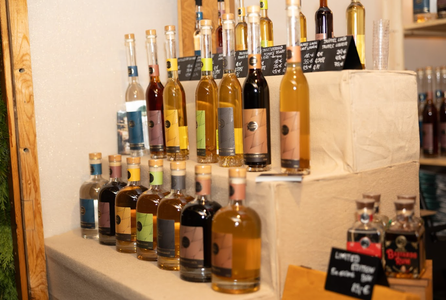A display of assorted bottled liqueurs on tiered shelves, each with colorful labels. Small chalkboard signs list prices. Warm, inviting atmosphere.