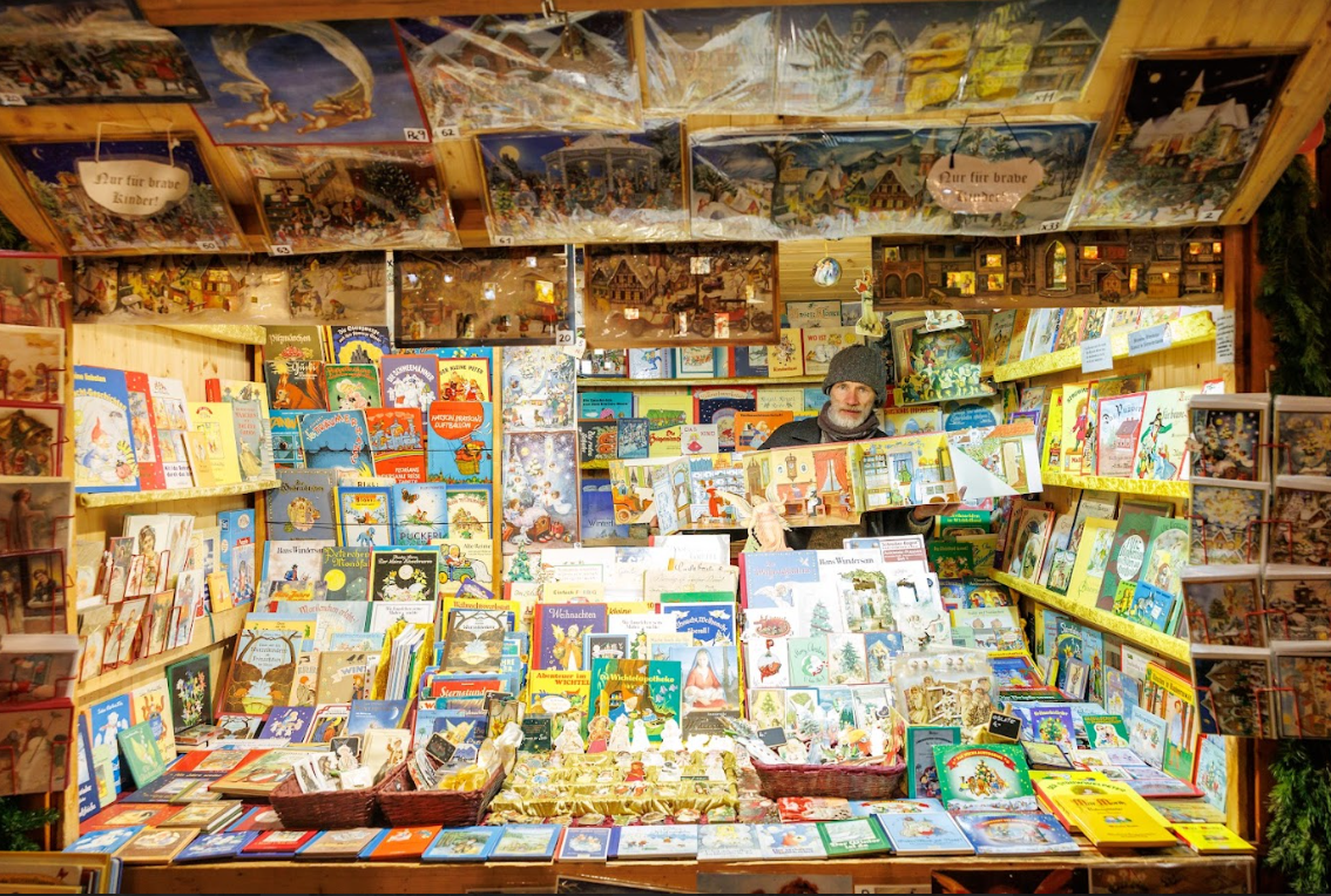 A festive market stall displays colorful children's books and storybooks, densely arranged. A vendor in a cozy hat smiles warmly behind the counter.