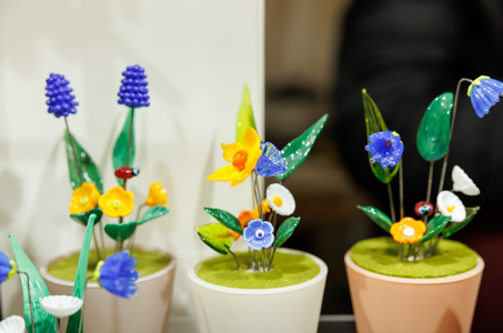 Three small potted displays feature vibrant glass flowers in shades of yellow, blue, and purple with green leaves, creating a cheerful, artistic scene.
