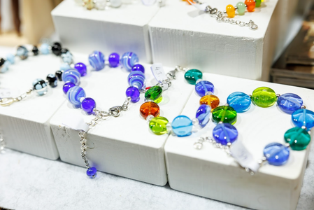 Colorful glass bead bracelets displayed on white blocks. The vibrant beads are in blue, green, orange, and purple, creating a lively, cheerful vibe.