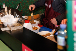 A person in a maroon apron serves food at a counter. A spatula is used to dish up a meal. Bowls, napkins, and bottles are visible on the counter.