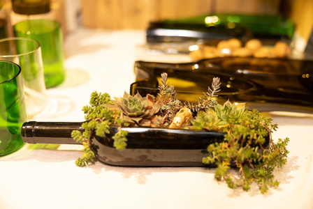 A wine bottle repurposed as a planter, filled with succulents and greenery, sits on a table beside green glasses and corks.
