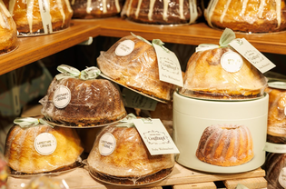 Assorted bundt cakes in clear wrapping are displayed on wooden shelves. Tags with green ribbons add a festive touch, conveying a cozy, inviting feel.