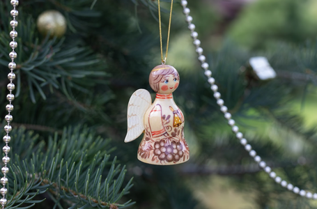 Wooden angel ornament with painted details hangs on a Christmas tree, adorned with beads and evergreen branches in the background.