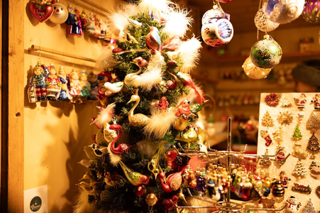 Festive market stall displaying a lush Christmas tree adorned with colorful, detailed ornaments. Nearby, hanging baubles and decorative pins add to the warm, joyful holiday atmosphere.