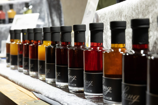 Row of small glass bottles filled with various colored liquids, each sealed with a black cap and labeled, displayed on a wooden shelf.