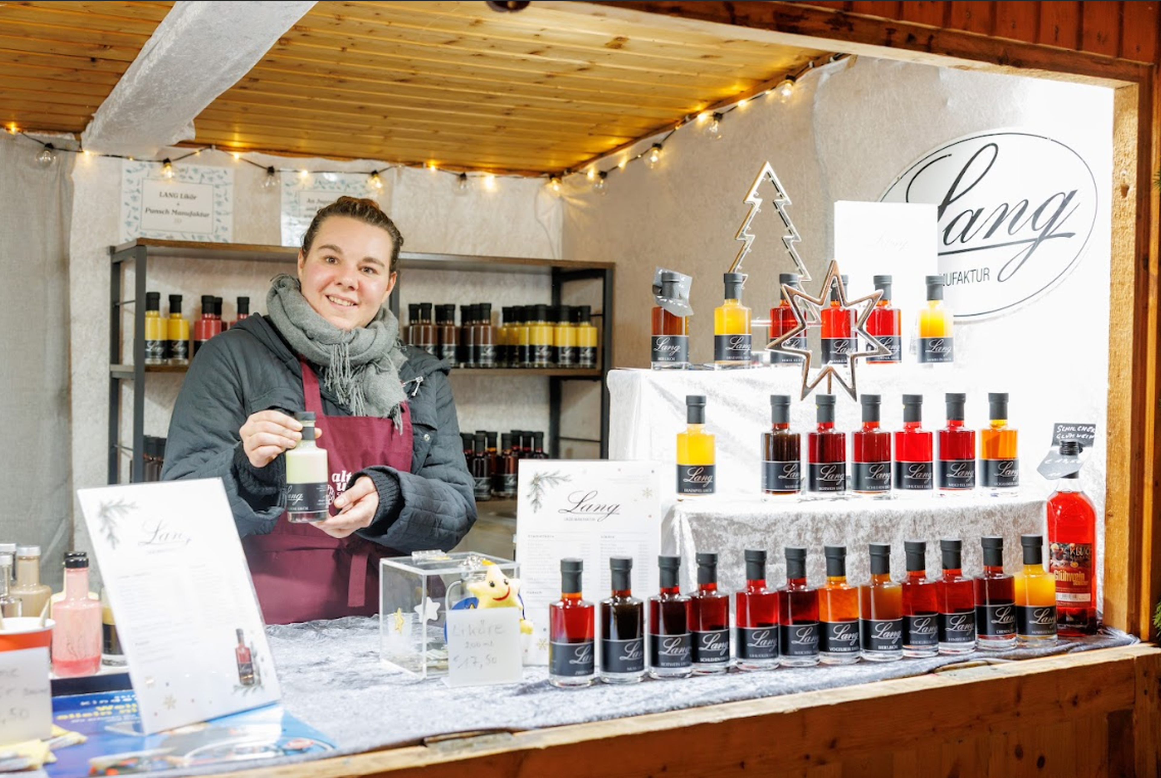 A smiling vendor in a cozy market stall displays colorful bottles arranged on tiered shelves. Warm lights create a festive, inviting atmosphere.