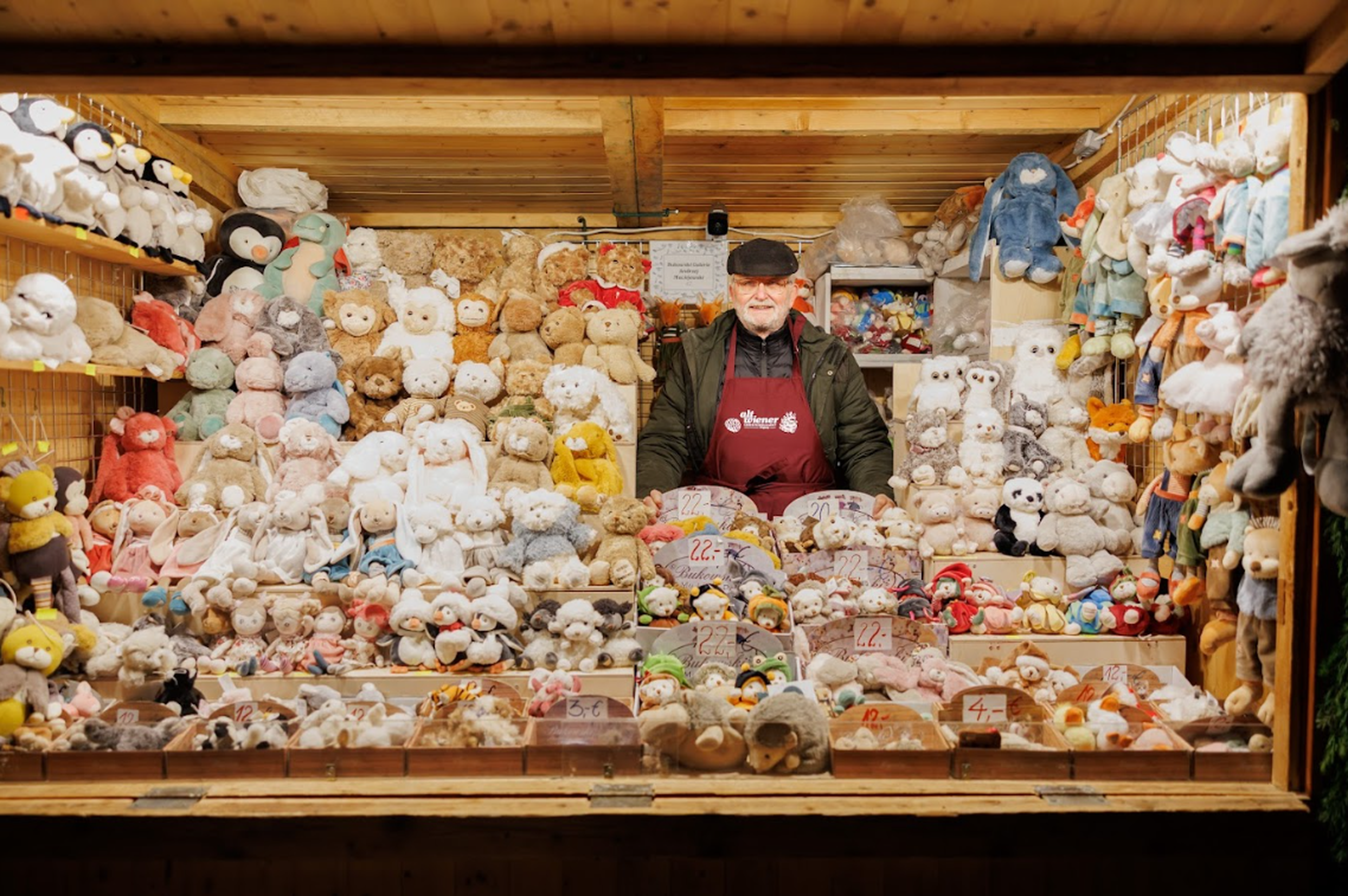 A vendor stands in a cozy wooden stall filled with colorful stuffed animals, displaying a cheerful and inviting atmosphere at a festive market.