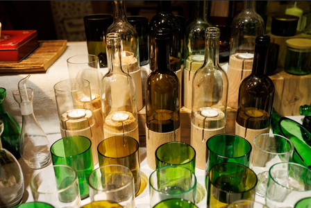 A variety of glass bottles and cups in green, amber, and transparent colors are creatively arranged with wooden holders on a table, evoking an artistic, cozy ambiance.