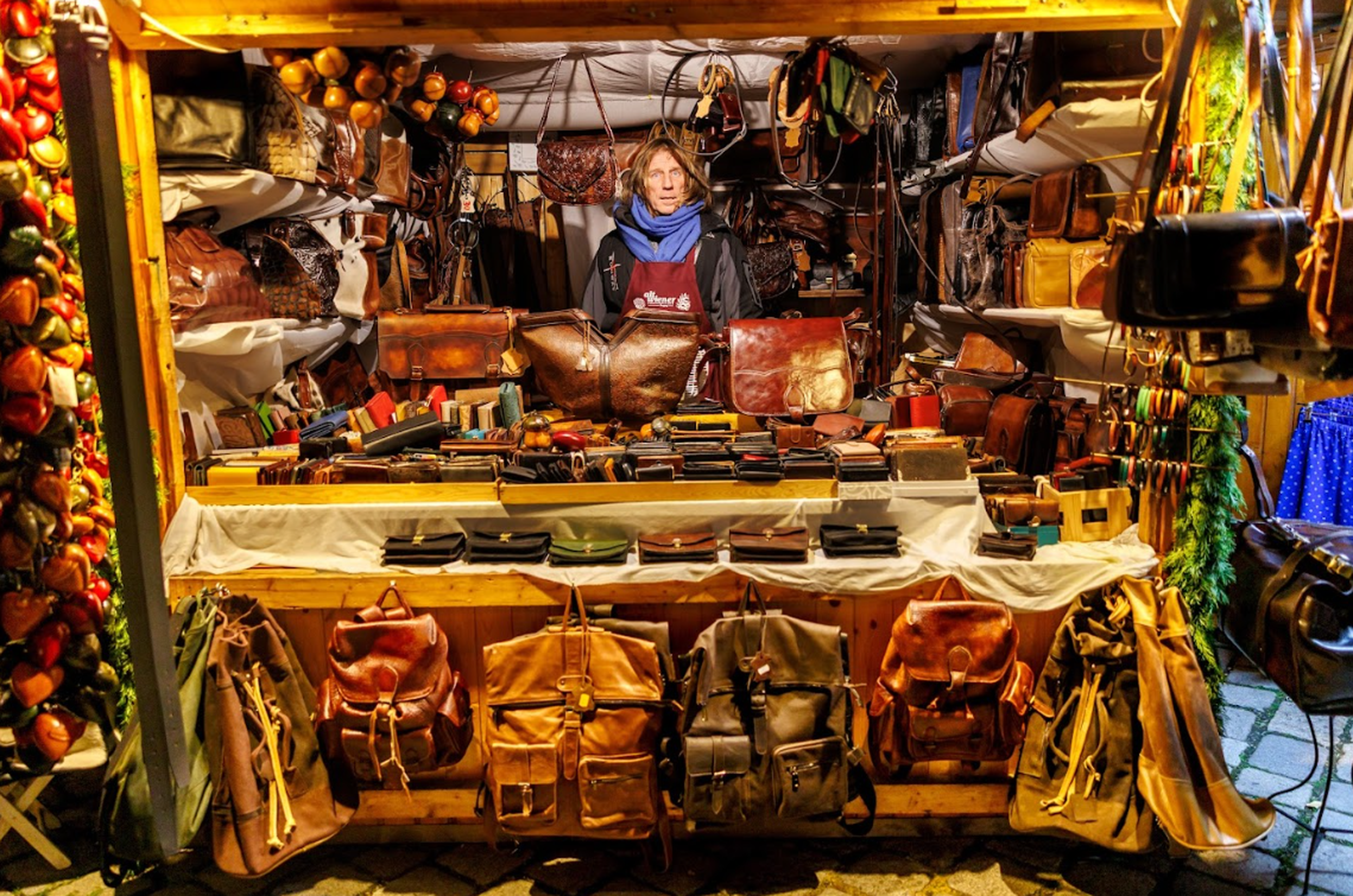 A vendor stands in a cozy, warmly lit stall filled with handcrafted leather goods. Bags and wallets in various shades are neatly displayed, creating an inviting atmosphere.