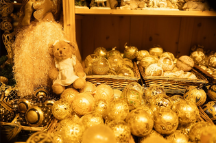 A teddy bear in a white dress sits among baskets of ornate, golden Christmas ornaments on a wooden shelf.