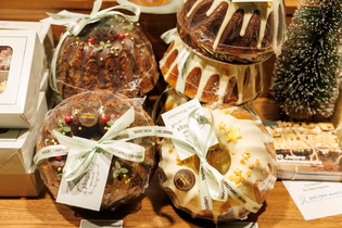 Assorted festive bundt cakes wrapped in plastic with ribbons, featuring chocolate and white icing with decorative candies. A small decorative tree is nearby.