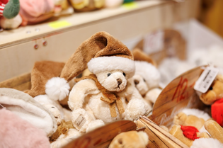 A plush teddy bear in a festive Santa hat and scarf is displayed on a shelf with other soft toys, conveying a cozy and cheerful holiday atmosphere.