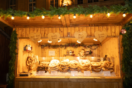 Wooden market stall with a smiling vendor, displaying piles of pretzels under warm lights. Festive, cozy atmosphere with greenery decorating the roof.