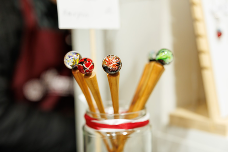 A glass jar holds wooden hair sticks topped with colorful, intricate glass marbles. The marbles are in red, green, and multicolored patterns.