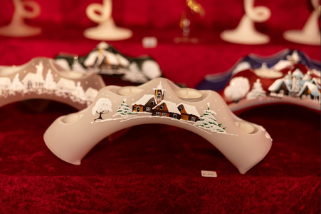 Decorative ceramic candle holder with a hand-painted winter village scene. Snow-covered houses and trees add a cozy, festive feel. Background displays similar items.