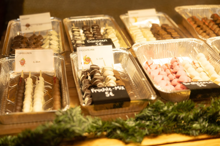 Trays of assorted chocolate-covered fruit skewers labeled with prices, displayed on a festive table with greenery.