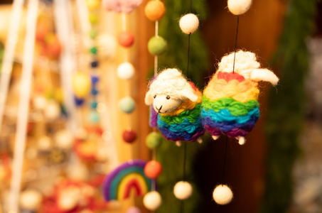Colorful yarn sheep ornaments with rainbow bodies and soft white faces hang amidst vibrant felt balls. A rainbow design softly blurs in the background.