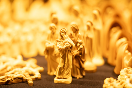 A warm-toned image of a nativity scene with detailed wooden figures; central figures depict a mother and child, surrounded by others in soft focus.