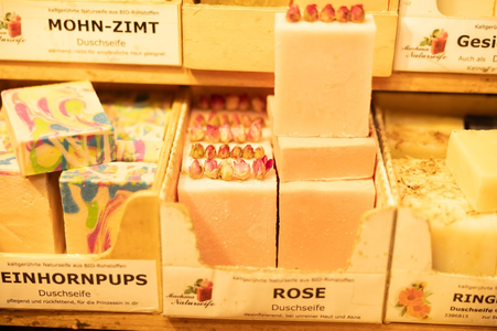 Display of colorful artisan soaps in wooden boxes. Varieties include a pink soap topped with dried roses, swirl-patterned, and cream-colored bars. Warm, inviting atmosphere.
