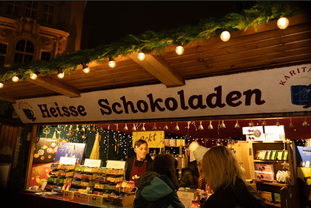 A festive market stall with a "Heisse Schokoladen" sign, selling hot chocolate and treats, adorned with lights and greenery.