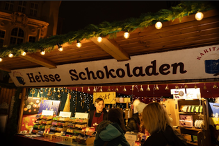 A festive market stall with a "Heisse Schokoladen" sign, selling hot chocolate and treats, adorned with lights and greenery.
