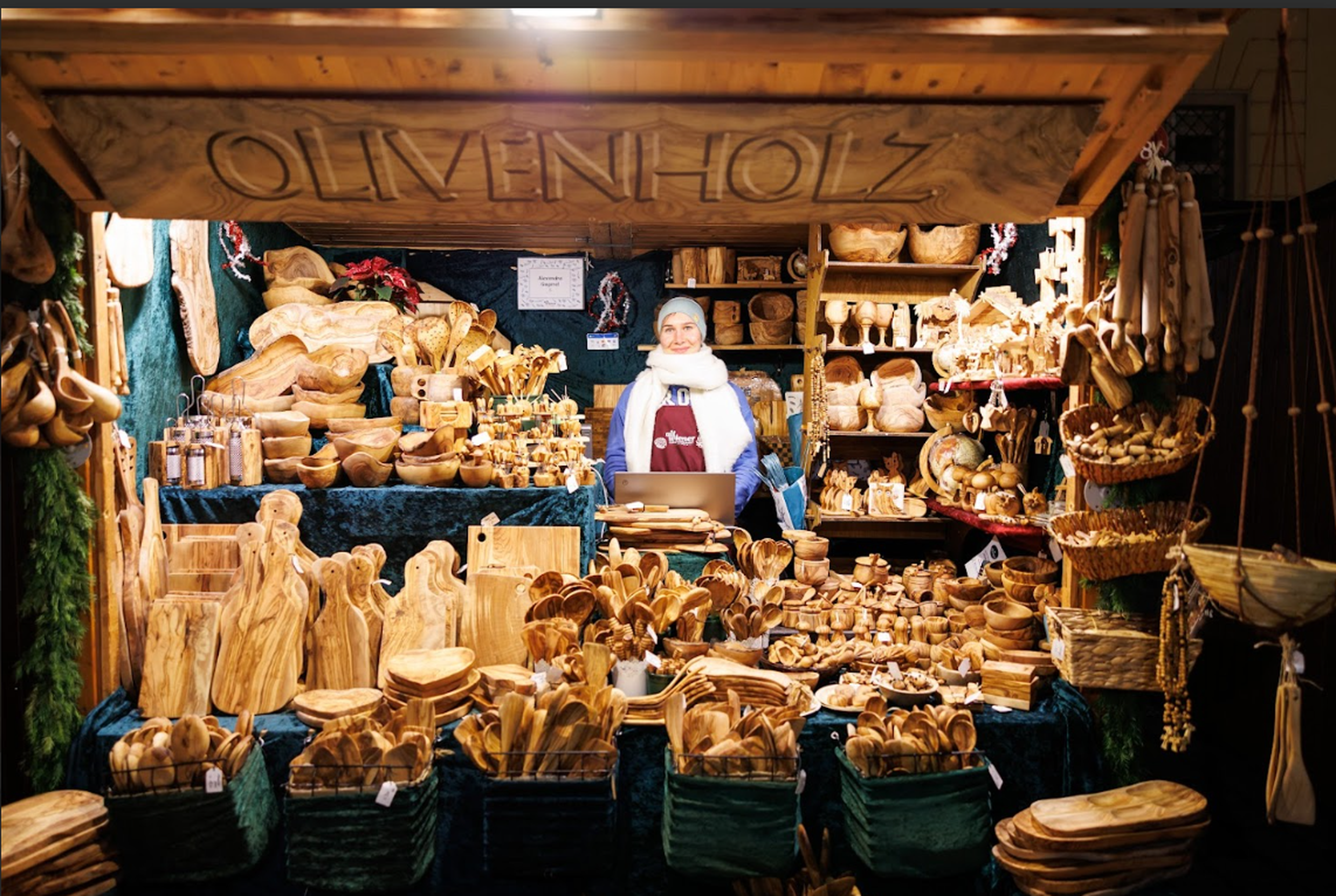 A vendor stands in a wooden crafts shop, surrounded by carved kitchen utensils, bowls, and decorative items on shelves and tables.