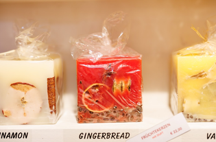 Three square candles with fruit inclusions are displayed; labeled cinnamon, gingerbread, and vanilla. Warm colors and a cozy vibe. Price is €22.90.