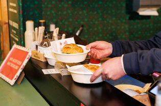 Hands holding bowls with semmel at a counter. A festive background and various items, including flyers and a sign, create a cozy atmosphere.