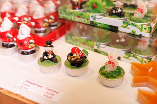 Small decorative tealights feature festive figures like Santas, snowmen, and lucky pigs with cheerful expressions, displayed on a shop counter.