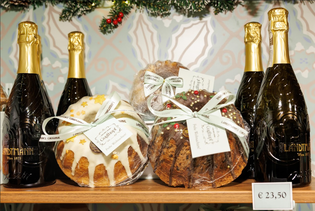 Bottles of sparkling wine and festive cakes topped with icing and embellishments are displayed on a wooden shelf. A price tag reads €23,50.