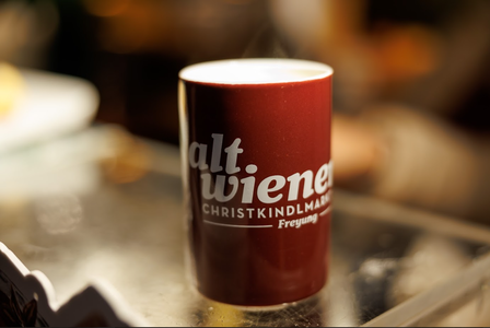 A steaming, maroon mug on a reflective surface, labeled "altwiener Christkindlmarkt Freyung," set against a blurred, warm-toned background.