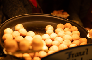 A large pan filled with golden, round doughnuts, warmly lit, with a blurred background of people holding trays.