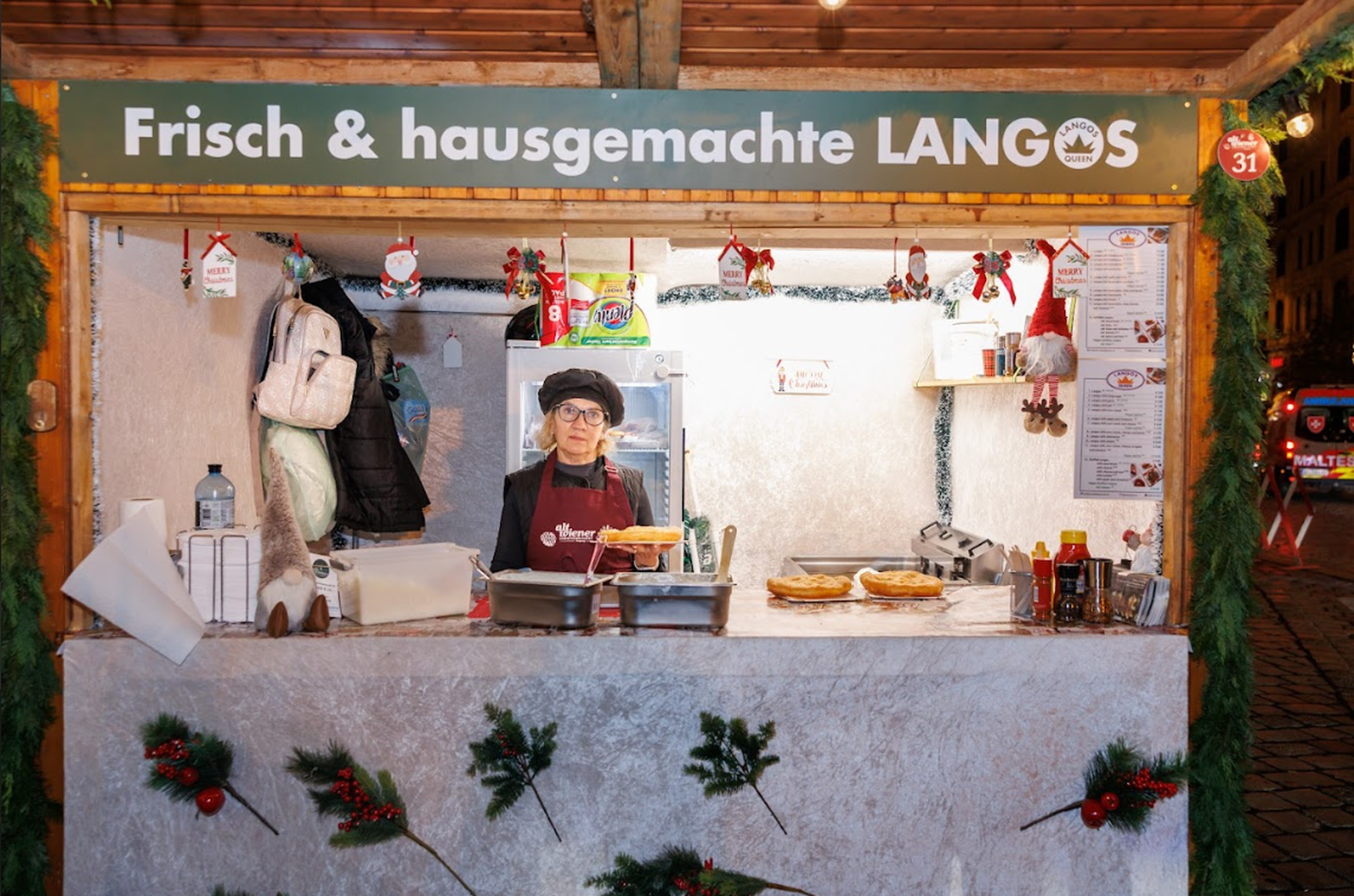 A cozy Christmas market stall with greenery and decor displays fresh homemade Langos. Inside, a person in a hat holds a Langos, exuding a welcoming atmosphere.