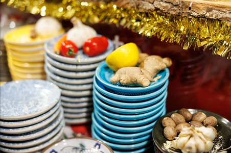 Stacks of colorful plates hold ginger, a lemon, garlic, and tomatoes. Nutmeg and cloves rest on a dish. Tinsel decorates the background, creating a festive atmosphere.